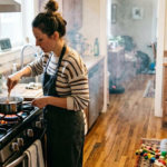 A mother cooking at the stove while her child plays on the ground in the background